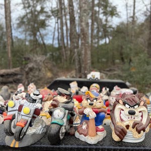 May include: A selection of Tazmanian Devil figurines, some on motorbikes, others with baseball bats or open-mouthed. The figurines are painted in various colours, including brown, red, and blue.