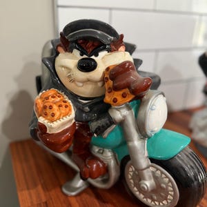 May include: A ceramic Taz cookie jar riding a teal motorcycle. Taz is wearing a black leather jacket and cap, holding a baked good. The motorcycle has silver accents and a black tyre. The cookie jar is on a wooden surface.