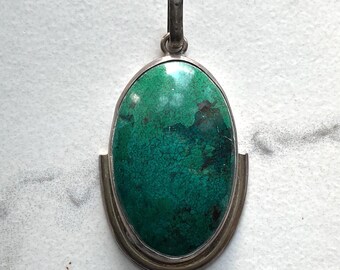 May include: Oval pendant with a vibrant teal stone set in a silver-toned metal frame. The stone has a textured surface with darker green and brown inclusions. The pendant has a loop at the top for a chain.
