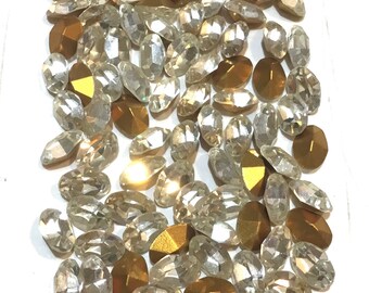 May include: A collection of clear and gold oval-shaped rhinestones. The rhinestones are scattered on a white surface.