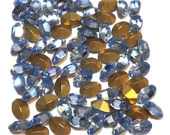 May include: A large assortment of light blue oval-shaped rhinestones with a iridescent finish. The rhinestones are scattered on a white background.