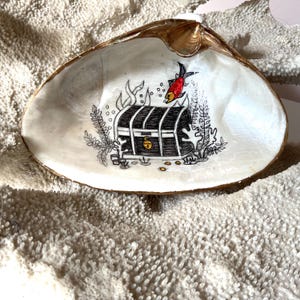 May include: A large, pearlescent seashell with a gold-painted rim. The interior features a hand-drawn illustration of a treasure chest, a red fish, and aquatic plants. The artwork is in black and white with gold accents.