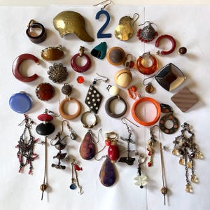 May include: A collection of 27 unique earrings in various colors, shapes, and materials. The earrings include wood, metal, plastic, and glass. Some earrings are simple and others are more elaborate. The earrings are arranged on a white surface. The number "2" is visible in the top center of the image.