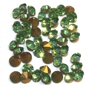 May include: A collection of green and gold rhinestones. The rhinestones are round and have a faceted surface. They are scattered on a white background.