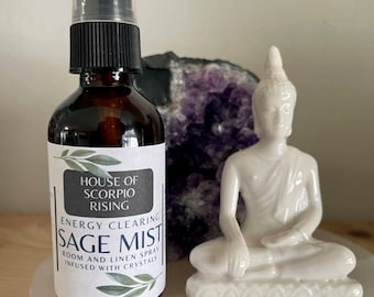 May include: A brown glass bottle with a black spray top labeled "House of Scorpio Rising Energy Clearing Sage Mist." A white ceramic Buddha statue sits next to the bottle. A purple geode is in the background.