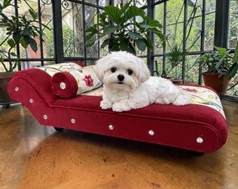May include: A small white dog rests on a red velvet pet chaise lounge. The chaise features a floral patterned cushion, crystal accents, and a matching bolster pillow. The dog bed is a luxurious item for pets.