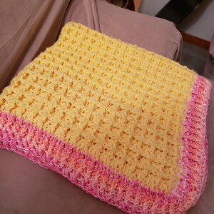 May include: A yellow crocheted baby blanket with a pink and orange crocheted border.