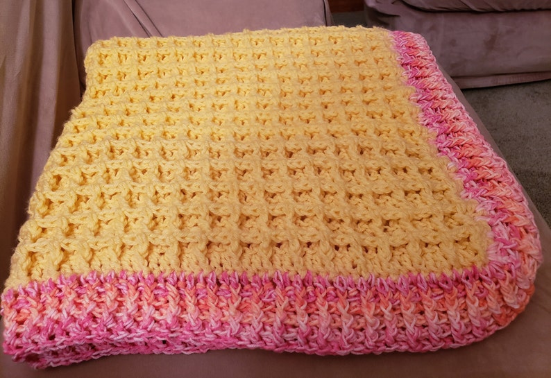 May include: A yellow crocheted baby blanket with a pink and orange crocheted border.