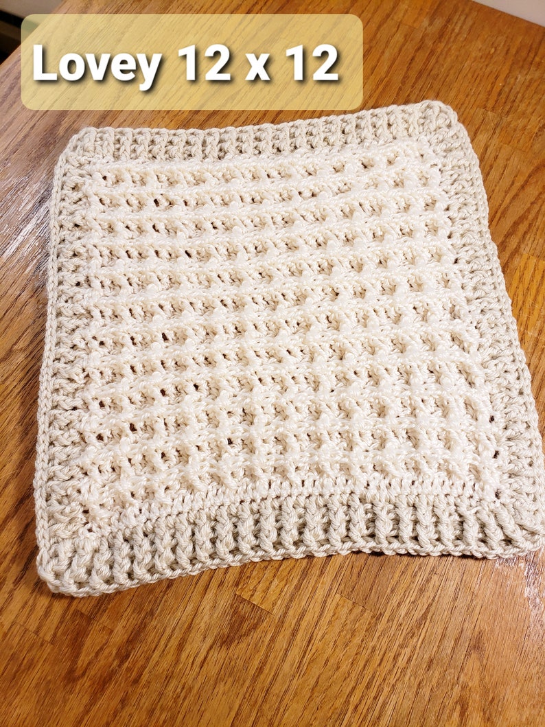 May include: A cream-colored crocheted square blanket with a textured pattern. The blanket is 12 inches by 12 inches and is labeled "Lovey 12 x 12".