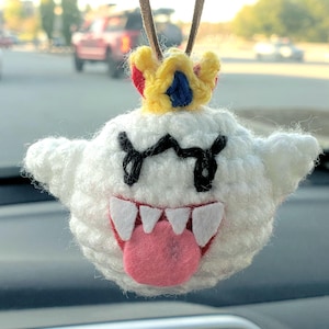 May include: A handmade white crochet ghost car charm with a yellow, red, and blue crown. The ghost has black eyes, a pink tongue, and white teeth. The charm hangs from a brown leather cord.