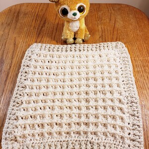 May include: A cream-colored crocheted blanket with a waffle stitch pattern. The blanket is lying flat on a wooden surface.