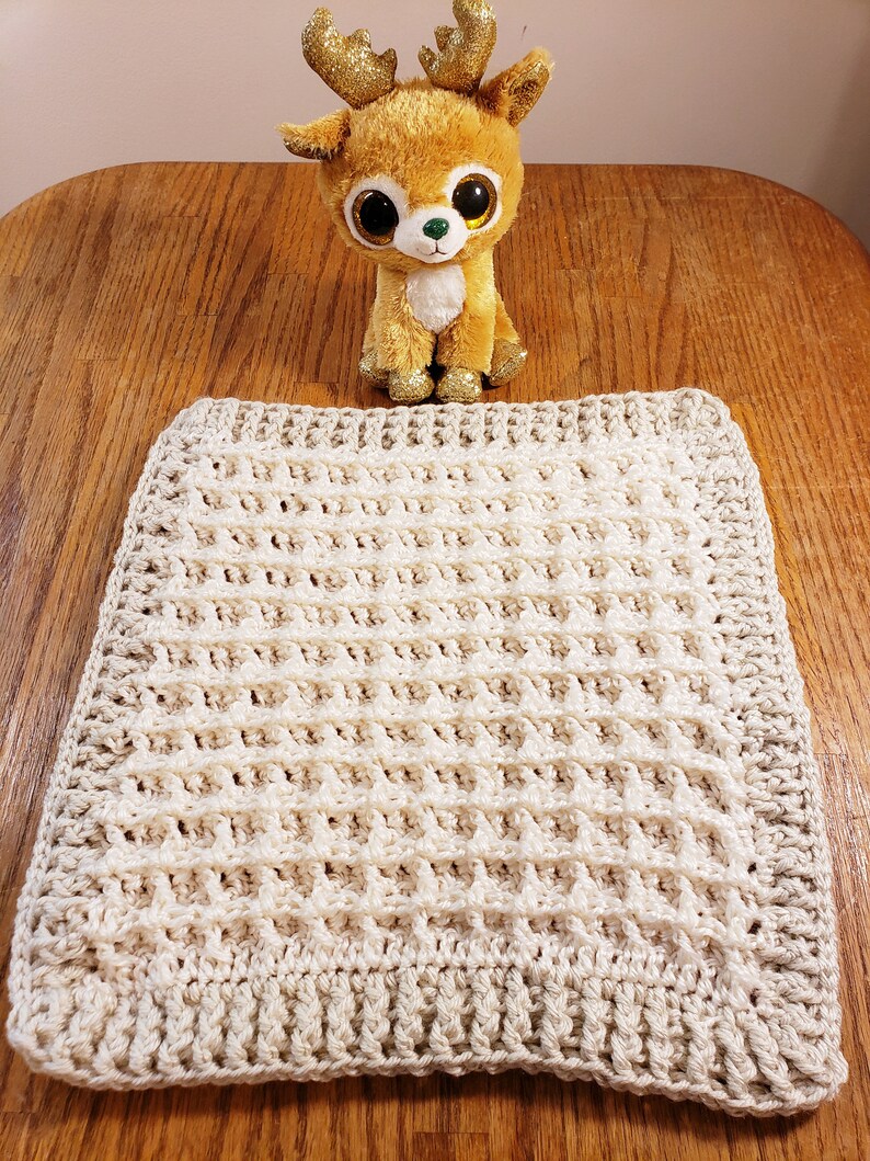 May include: A cream-colored crocheted blanket with a waffle stitch pattern. The blanket is lying flat on a wooden surface.