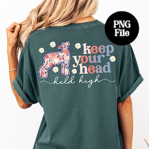 May include: Dark teal t-shirt with a floral lamb graphic and the text "keep your head held high." The lamb is filled with a colorful floral pattern. White daisy accents are scattered around the text and lamb.