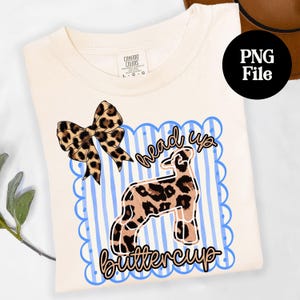 May include: Cream-colored t-shirt with a leopard print bow and a graphic of a lamb with leopard print spots. The text "head up buttercup" is written in cursive. The shirt has a Comfort Colors tag.