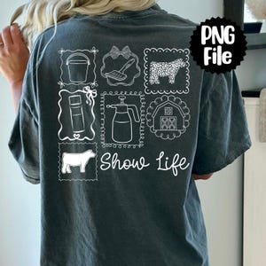 Puede incluir: Camiseta gris oscuro con un diseño gráfico blanco que presenta imágenes relacionadas con la agricultura y el texto "Show Life". El diseño incluye un cubo, un lazo, un cepillo, una vaca, un pulverizador, un granero y una vaca. La camiseta es de estilo casual y cómodo.