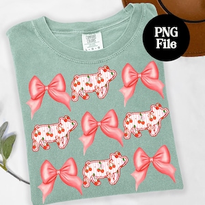 May include: A light green t-shirt featuring a pattern of pink bows and pig illustrations. The pigs have a cherry design and are outlined in red. The shirt has a Comfort Colors tag.