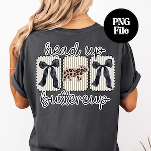 May include: Dark gray t-shirt with the words "head up buttercup" in a script font. The design includes three decorative elements: two black bows and a pig with a leopard print pattern.