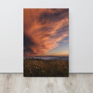 May include: A vertical canvas print featuring a landscape scene at sunset. The sky is filled with dramatic, swirling orange and pink clouds above a dark, silhouetted mountain range. In the foreground, a field of tall, wild grasses is visible.