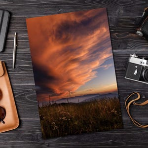 May include: A rectangular print featuring a landscape photograph of a grassy field under a dramatic sunset sky with orange and purple clouds. The print is displayed on a dark wooden surface alongside a silver pen, a camera, and a leather case.
