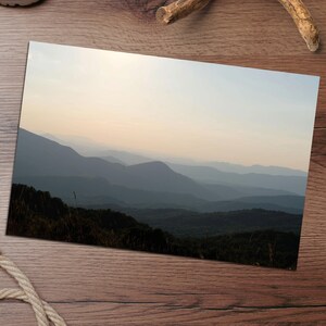May include: A rectangular photographic print featuring a landscape of layered mountain ranges fading into a soft, pale sky at dusk. The foreground shows dark, silhouetted hills covered in dense vegetation, contrasting with the light, hazy atmosphere.