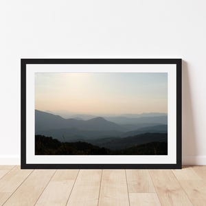 May include: A framed landscape photograph featuring layers of blue-toned mountain silhouettes fading into a soft, pale sky at sunrise or sunset. The image is enclosed in a simple black frame with a white mat, displaying a serene mountain vista.
