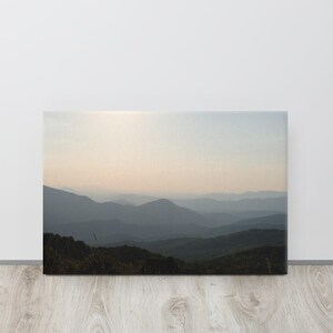 May include: A rectangular canvas print featuring a landscape of layered mountain ranges fading into the distance. The foreground shows dark, silhouetted vegetation, while the sky above displays a soft, pale gradient of light during sunrise or sunset.