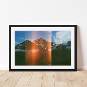 May include: A framed landscape photograph featuring a mountain range under a blue sky with clouds. A prominent vertical orange light leak effect runs through the center of the image, casting a warm glow over the dark green forested slopes.