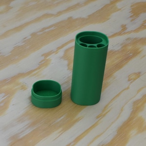 May include: A green cylindrical container with a lid. The container has multiple compartments inside. The container is on a light brown wooden surface.
