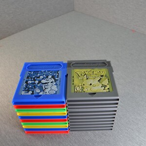May include: Two stacks of Game Boy cartridges. One stack is blue with a Super Mario Bros. graphic, and the other is gray with a Pokémon Pikachu Edition graphic. The blue stack has a rainbow-colored edge.