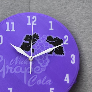 Puede incluir: Un reloj de pared morado con forma de tapón de botella, con números y manecillas blancos. La esfera del reloj muestra el texto "Nuk Grape Cola" y un diseño de uvas. El reloj está sobre un fondo gris.