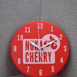 Puede incluir: Reloj de pared rojo con números y manecillas blancas. La esfera del reloj presenta el texto "Nuka Cherry" en blanco, con un logotipo de cereza estilizado. El reloj tiene un diseño de tapón de botella.