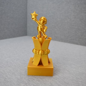 May include: A gold-colored 3D printed trophy featuring a character holding a star. The trophy has a square base and a multi-tiered design.