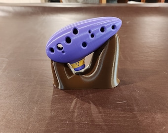 May include: A purple ocarina with a stand. The ocarina has eight finger holes and a yellow and gold design on the bottom. The stand is brown and is made of plastic.