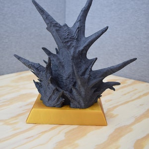 Könnte beinhalten: Eine schwarze, stachelige, abstrakte Skulptur auf einem goldenen Sockel. Die Skulptur ist aus einem Material gefertigt, das wie Holz oder Stein aussieht.