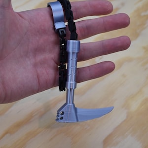 Puede incluir: Un cuchillo de dedo plateado con una cadena negra y una hoja dentada. El cuchillo se sostiene en una mano sobre un fondo de vetas de madera. El cuchillo de dedo es una herramienta única.