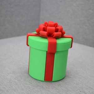 Könnte beinhalten: Eine grüne, zylindrische Geschenkbox mit rotem Band und Schleife. Die Box ist etwa 10 cm hoch und 8 cm im Durchmesser. Das rote Band umschließt die Box und ist mit einer großen Schleife verziert.