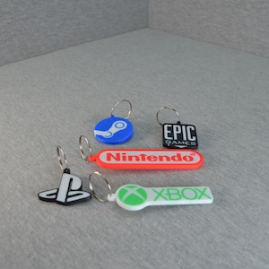 Puede incluir: Varios llaveros con temática de videojuegos. Incluyen llaveros con los logotipos de Steam, PlayStation, Epic Games, Nintendo y Xbox. Los llaveros son de varios colores, como azul, rojo, negro y blanco. Cada uno tiene un anillo de metal.