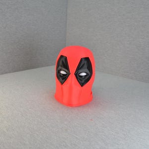 May include: A bright red mask with black eye and mouth details, reminiscent of a comic book character. The mask is a solid color with a smooth texture, and the eye shapes are outlined in black. The mask is a pop culture item.