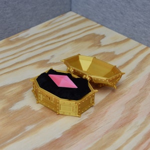 May include: A gold-colored coffin-shaped box, open to reveal a pink, geometric gem resting on black velvet. The box has decorative rivets and is set on a wooden surface.