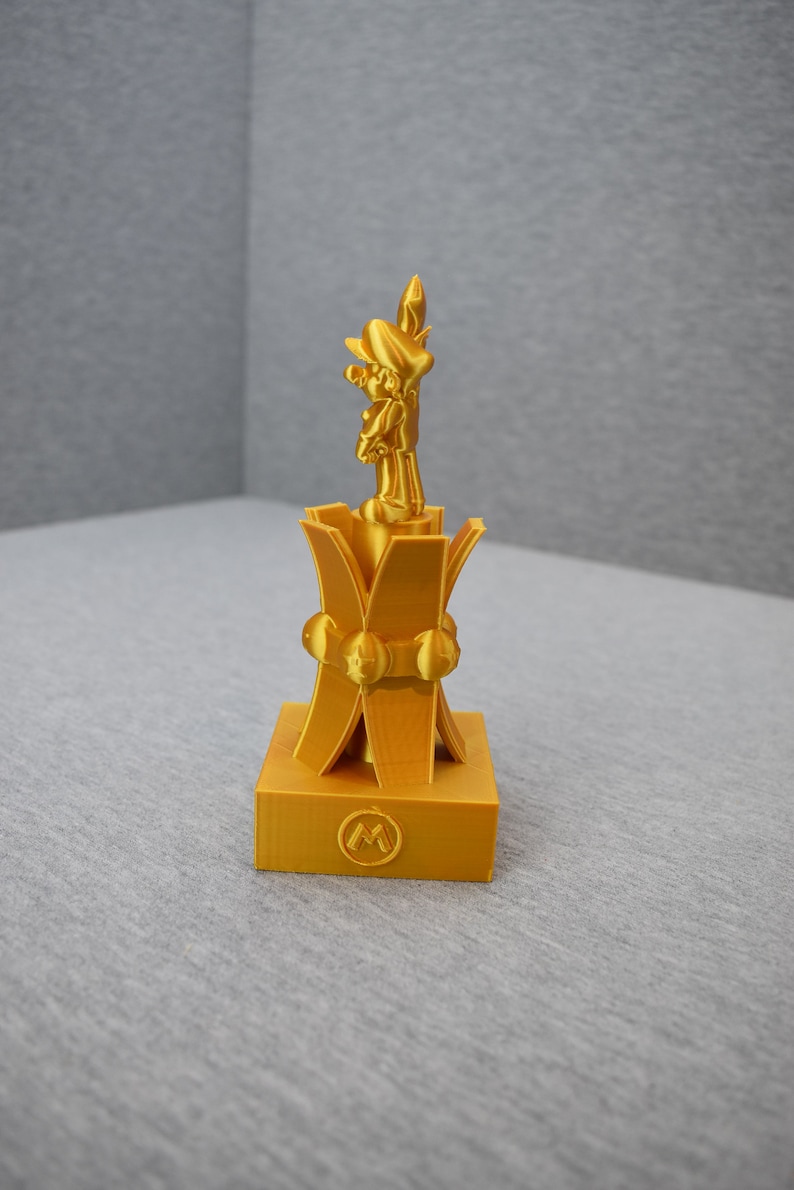May include: A gold-colored 3D printed trophy featuring a character from the Super Mario franchise standing on a base with a stylized "M" logo.