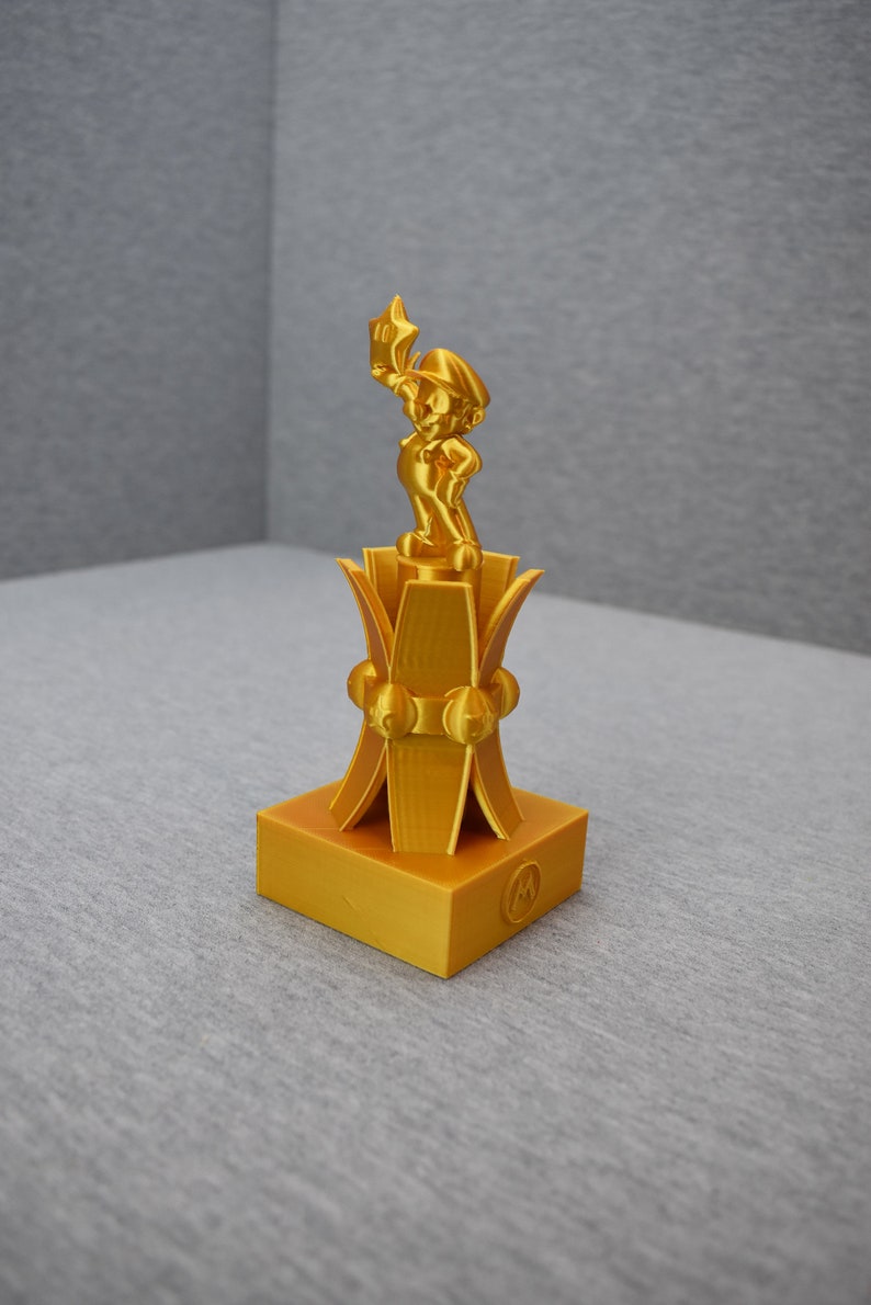 May include: A gold-colored 3D printed trophy featuring a figurine of a video game character holding a star. The trophy has a base with a stylized design and a logo.