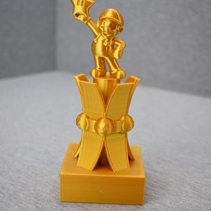 May include: A gold-colored 3D printed trophy featuring a character holding a star. The trophy has a square base and a multi-tiered design.