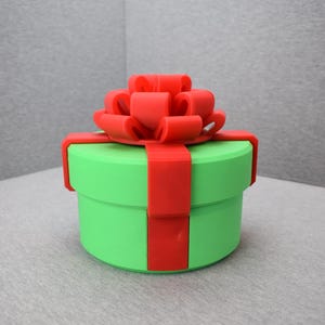 Könnte beinhalten: Eine runde, grüne Geschenkbox mit einem leuchtend roten Band und einer Schleife. Die Box hat einen Durchmesser von etwa 10 cm und ist mit einem passenden roten Band umwickelt. Die Schleife sitzt oben auf der Box.