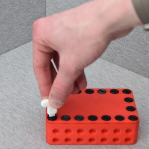 Op de afbeelding: Een rode plastic doos met zwarte cirkels rond de randen en kleinere cirkels in de hele doos. Een wit plastic stuk wordt in een van de grotere cirkels gedrukt.