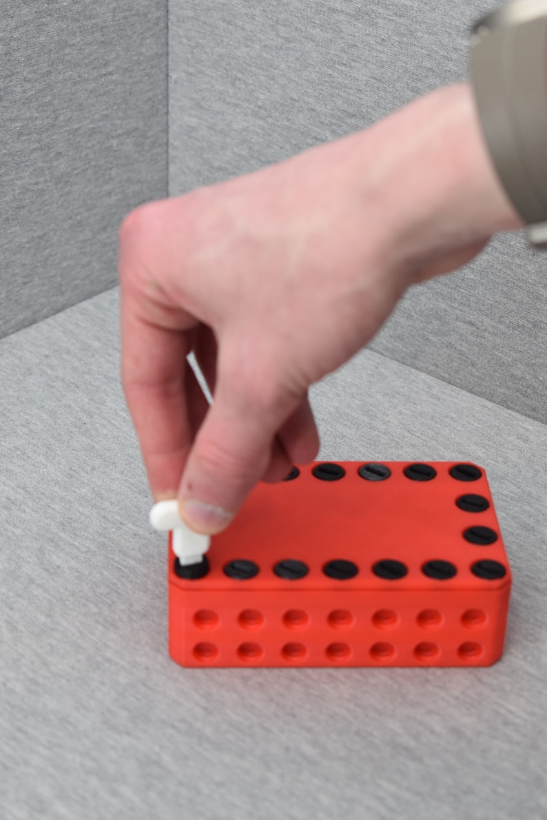 Op de afbeelding: Een rode plastic doos met zwarte cirkels rond de randen en kleinere cirkels in de hele doos. Een wit plastic stuk wordt in een van de grotere cirkels gedrukt.