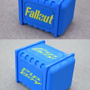 以下が含まれることがあります： 上面が黄色い「Fallout」ロゴ、側面が黄色い「Pip-Boy」ロゴの青いプラスチック製の箱です。