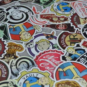 May include: A collection of colorful stickers featuring various designs and logos. The stickers include images of a cartoon character, bottle caps, and the text "Nuka Cola", "Vault-Tec", and "Red Rocket".