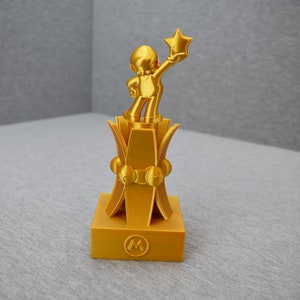 May include: A gold-colored trophy featuring a cartoon character holding a star. The trophy has a square base with a circular "M" logo.