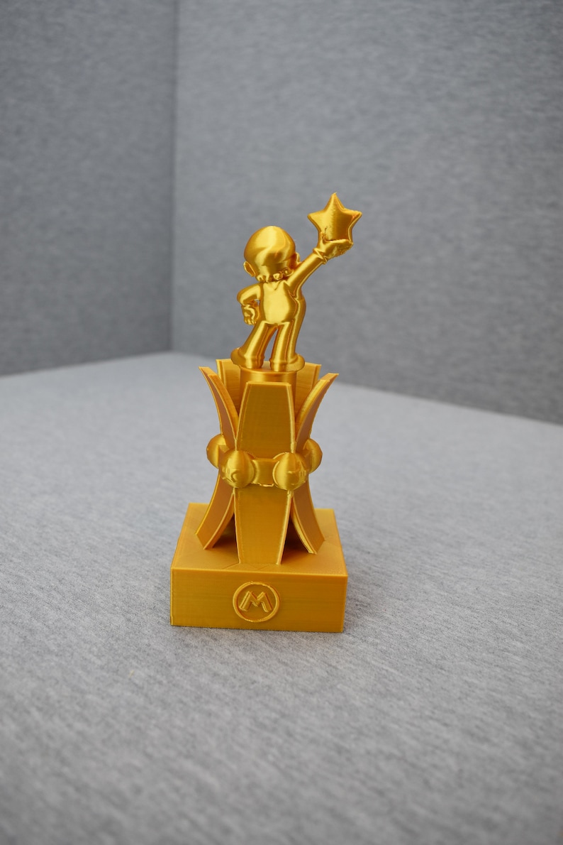 May include: A gold-colored trophy featuring a cartoon character holding a star. The trophy has a square base with a circular "M" logo.