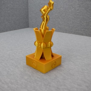 May include: A gold-colored trophy with a stylized figure on top. The base of the trophy is square with a decorative design.